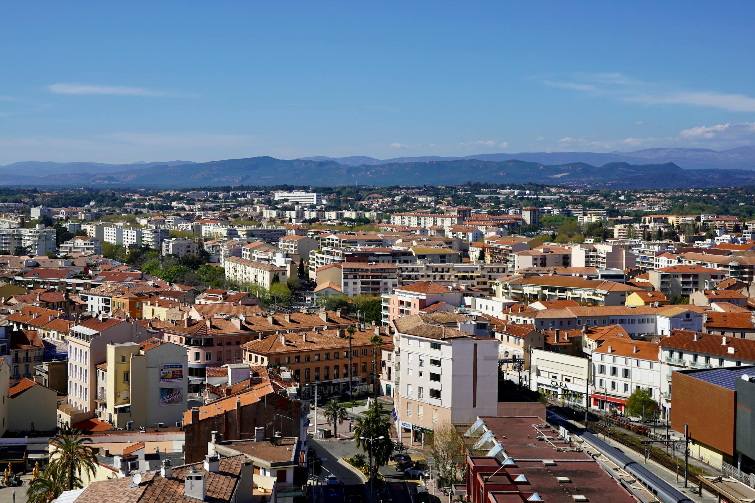 Aerial view of a Mediterranean city with mountains in the background under a clear blue sky.