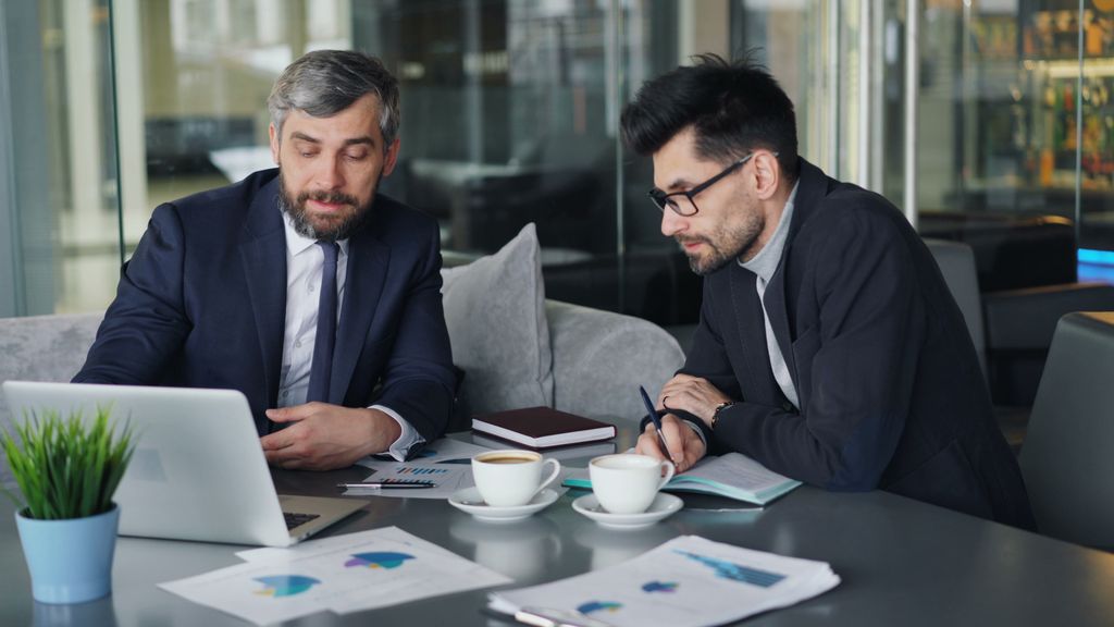 Two businessmen in suits reviewing charts and graphs over coffee in a modern office setting.